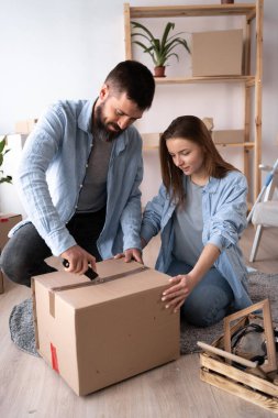 Taşındığı gün bir erkek ve bir kadın yere oturup eşyaları boşaltıyorlar. Adamın biri karton bir kutunun üzerindeki bandı bıçakla kesiyor. Yeni bir dairede mutlu bir çift. Boşluğu kopyala.