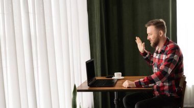 Milenyum boyunca günlük giysiler içindeki beyaz adam evde bir masada dizüstü bilgisayar kullanarak otururken video görüşmesi yapıyor. Merhaba demek için elini kaldırdı. Pankart. Boşluğu kopyala.