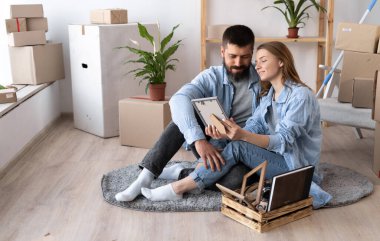 Mutlu genç çift, rahat giysiler içinde yerde oturup çerçevelenmiş fotoğrafa sarılıyorlar. Beyaz bir duvarın yanında karton kutular ve bitkiler yığını. Taşınma günü. Boşluğu kopyala.