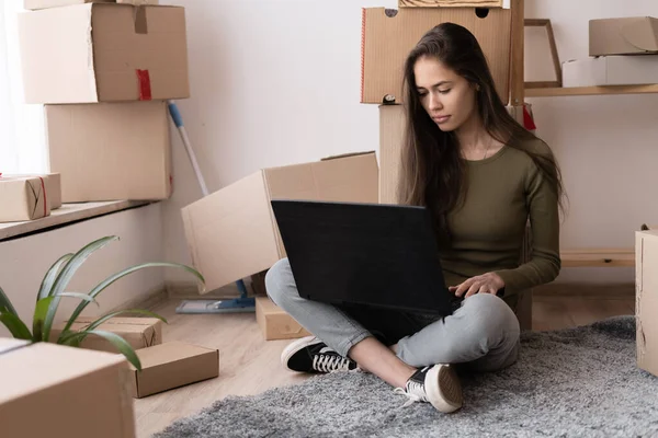 happy girl with boxes moves to new apartment, moving to a new house, rental housing, spanish young woman using laptop computer to search and order a transportation service and movers to move to a new home