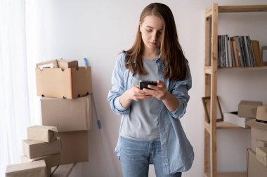 Ciddi genç bir kadın taşınıyor, karton kutuların arasında duruyor, akıllı telefon kullanıyor, yeni bir eve taşınıyor, taşınma günü ve teslimat çağrısı, daireye çevrimiçi sipariş,