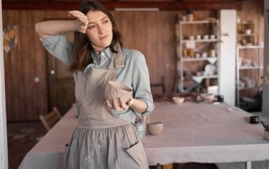 Çamurdan bir kase yontmaya odaklanmış yorgun genç kadın çömlekçi çömlekçilik dersi ya da atölye çalışması zamanı, yaratıcı bir stüdyoda bir kadın. Alnından terini elle sil..