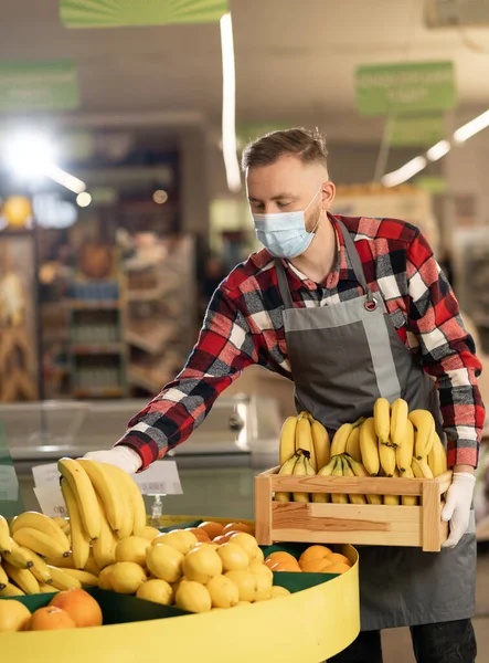 male grocery store worker wearing face shield and apron standing in store and sorting fruit. Handsome caucasian salesman in mask working indoors. Supermarket concept