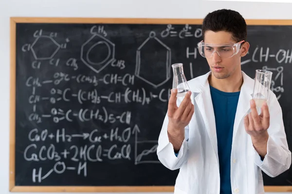 Man chemist student with glass flasks in classroom. Young arabic scientist holding laboratory flask on blackboard background, science and education concept — 스톡 사진