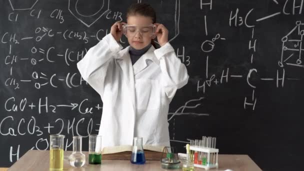 Een kleine vrouwelijke wetenschapper in een witte jas en bril onderzoekt een reageerbuis met een chemisch reagens. Onderzoek en onderwijs op school. — Stockvideo