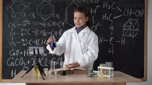 Ittle scientist girl in white coat leads an online video blog, records video using smartphone. Research and education. Chemical laboratory with glass flasks. Schoolgirl conducts a chemical experiment — 图库视频影像