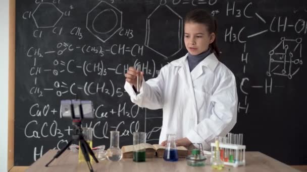 Little scientist girl in white coat leads an online video blog, records video using smartphone. Research and education. Chemical laboratory with glass flasks. Schoolgirl conducts a chemical experiment — Stockvideo