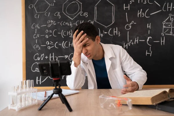 Upset online teacher giving chemistry lesson using smartphone, arabic tutor or student sitting at table with cell phone, chemistry online video conference, school webinar, online education concept
