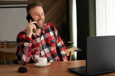 Caucasian smiling man in red shirt using laptop for online shopping, talking on mobile phone and drinking coffee in coffee shop while working as online freelancer