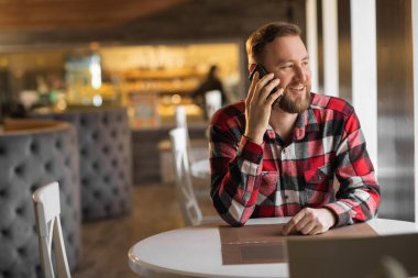Kafeden çalışıyorum. Gündelik giyinen genç adam yaratıcı kafenin penceresinin yanında otururken cep telefonuyla konuşuyor. Hafta sonları kafede çalışan bir adam. teknoloji iletişim konsepti