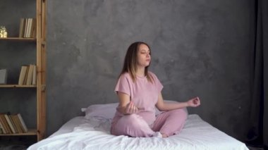 Genç hamile bir kadın, evde uyandıktan sonra yoga egzersizi yapıyor. Yogi kadın modeli yatakta bağdaş kurup meditasyon yapıyor, hamilelik sigortası yaptırıyor.