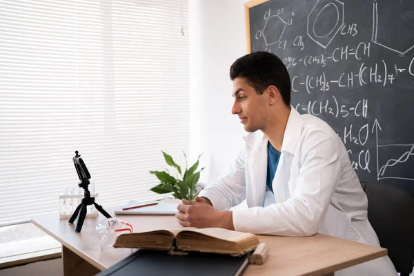 Modern day teacher. young muslim male tutor explaining learning material before smartphone webcam. Confident arabic man chemistry coach broadcasting live training online speaking to students