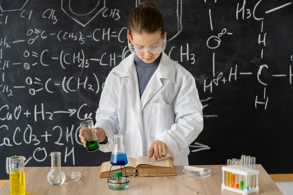 young adorable student girl writing a chemistry result on book while doing a scientific experiment and standing in the modern laboratory with chalkboard as background. Early development of children.