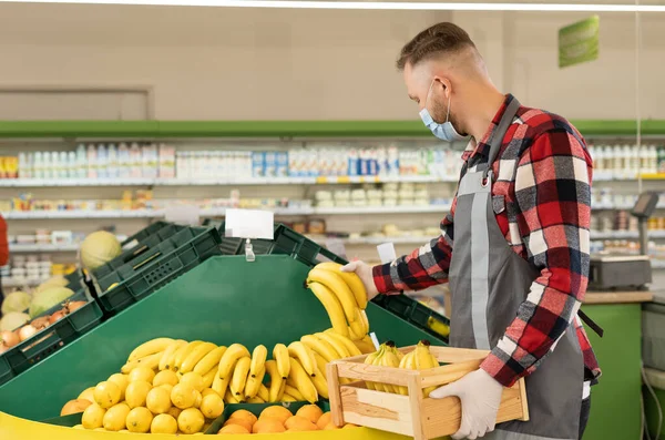 Caucasian employee in fruits store with citrus and bananas, supermarket worker in a protective face mask putting bananas out of a box on store shelves, organic product concept