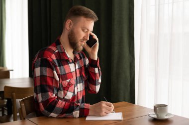 Kahve molası sırasında cep telefonuyla konuşan, not defterine bilgi yazan, operatörle telefon görüşmeleri sırasında not alan, serbest çalışan, teknolojiyi kullanan gülümseyen adam.