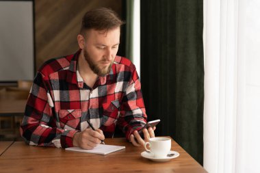 Dijital öğrenme, çevrimiçi çalışma, akıllı telefon ve kağıt not defterine yazı yazan sıradan adam ya da öğrenci, kafede ya da iç mekanda cep telefonu kullanan genç adam, çevrimiçi çalışıyor.