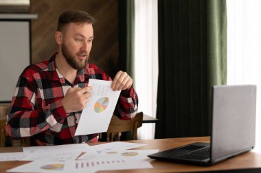 Sanal buluşma konsepti. Bilgisayarlı kameraya diyagramlı belgeleri gösteren genç bir adamın ofiste çalışırken tartıştığına eminim.