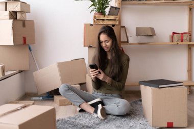 Yeni evin oturma odasında akıllı telefon kullanan mutlu, genç bir İspanyol kadın taşınma gününde bir yığın karton kutuyla, gülümsüyor.