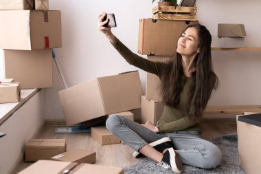 Mutlu esmer kız akıllı telefonuyla selfie çekiyor ve evde karton kutuların arasında oturuyor, yeni ev, teknoloji, insanlar, yenileme ve yer değiştirme konsepti