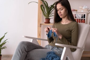 Latin genç kadın evde koltukta otururken örgü örüyor, hobi ve dikiş işi, el yapımı örgü, boş zaman ve hobi konsepti.