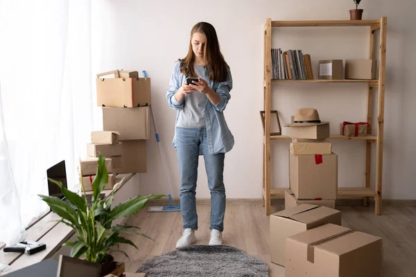 新家的客厅里 一个快乐的年轻女人在搬家的那天用智能手机和一堆纸板箱在一起 — 图库照片