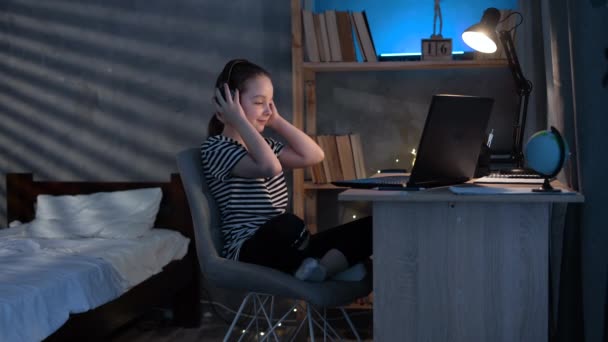 带着笔记本电脑的小女孩深夜在家里听音乐 晚上带着耳机的孩子在餐桌边休息 — 图库视频影像