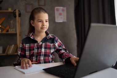 Küçük kız öğrenci evde oturmuş dizüstü bilgisayara bakıyor notlar alıyor, beyaz kız bilgisayarda webinar izliyor, evde eğitim görüyor. uzak eğitim kavramı.