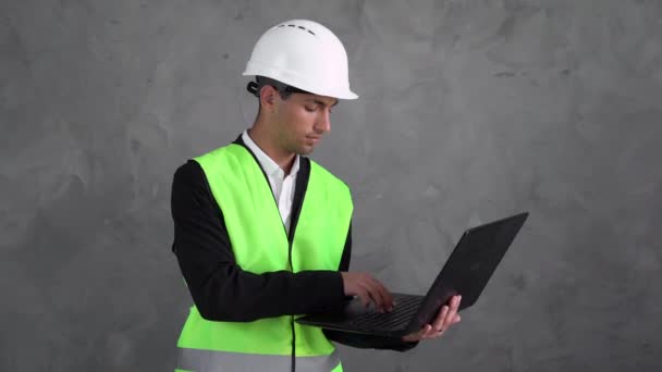 年轻的男性建筑师 穿着白色头盔 在笔记本电脑上发短信 进行商务交流 一个信任的微笑看着摄像机 穆斯林工头复制空间 — 图库视频影像