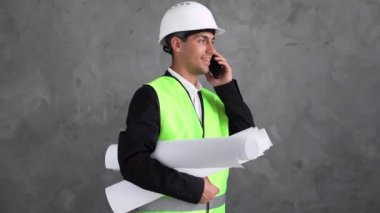 Aktif, mutlu Müslüman, mimar, inşaat işçisi, elinde gri arka planda izole edilmiş bir binanın mimari planları var. Mühendis, akıllı telefondan müşteriye telefon ediyor. İnşaat