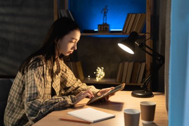 Asyalı kız öğrenci akşamları masa başında otururken dijital tableti kullanıyor.