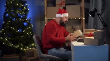 Takım elbiseli mutlu idus sakallı Noel Baba, evdeki bir ofiste, kutuların arasında oturuyor, dizüstü bilgisayarına envanter notları yazıyor. Noel teslimatı. Portre.