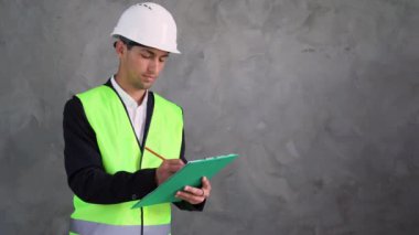 Arap yakışıklı mühendis ya da mimar, beton bir duvarın arka planında duruyor, elinde bir tablet ve beyaz kağıt üzerinde bir kalemle notlar tutuyor. Beyaz bir şapkayla çalışan bir adam.