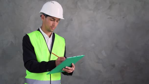 阿拉伯英俊的工程师或建筑师站在水泥墙的后面 手里拿着一块石碑 手里拿着一支笔在白纸上写字 头戴白色硬礼帽的工人 — 图库视频影像