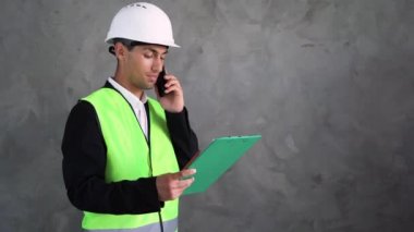 Genç bir Arap erkek mimar, beton bir duvarın arkasında beyaz bir miğferin içinde duruyor, akıllı bir telefonla konuşuyor ve tabletten bir şeyler okuyor. Boşluğu kopyala.