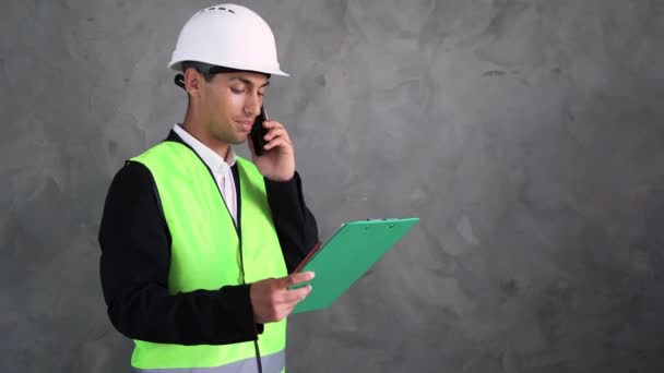 一位年轻的阿拉伯男性建筑师头戴白色头盔 站在水泥墙的后面 一边用智能手机说话 一边在平板电脑上看书 复制空间 — 图库视频影像