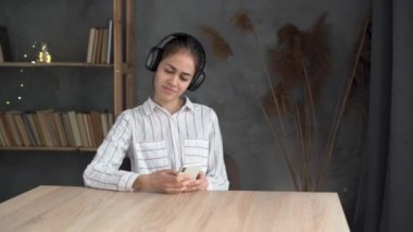 Huzurlu genç bir kadın, evde rahat rahat oturuyor, günlük kıyafetlerini giyiyor, kablosuz kulaklık takıyor, hafta sonunun keyfini çıkarıyor, cep telefonunda internet uygulaması kullanıyor, müzik dinliyor, kapalı gözler kullanıyor.