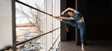 Hispanic athletic woman doing leg stretching exercise in the gym standing by the window, fitness and stretching twine concept. banner, place for text