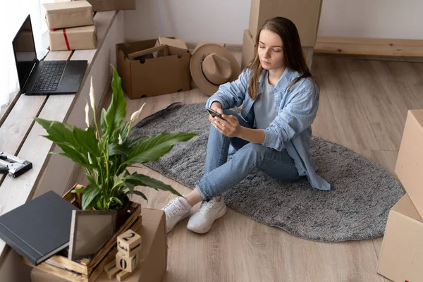 Beautiful girl is using a smartphone, looking at camera and smiling while sitting on the floor near packed boxes ready to move, young woman texting while moving to new apartment. top view
