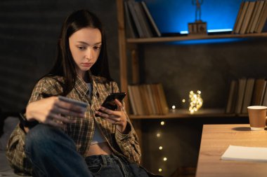 Asyalı kız kredi kartı tutuyor ve gece evde telefon kullanıyor. Çevrimiçi alışveriş, E-ticaret, İnternet bankacılığı, para harcama kavramı