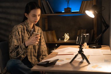 Asyalı kız öğrenci gece geç saatlerde evde akıllı telefon kullanarak ders çalışıyor, bayan öğrenci kahve içiyor ve akşam sınava hazırlanıyor.