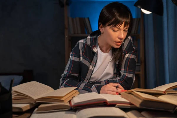 Young woman studying at home in the evening sitting at a table with books, home education or teacher preparing for a lesson, teaching at night concept