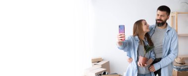 Yeni bir dairede selfie çeken genç bir çift, aydınlık eve aile taşınması, akıllı telefonlu bir adam ve kadın, yeni ev hamlesi konsepti, hafıza fotoğrafı, afiş