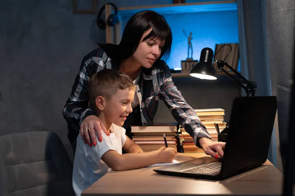 Young mother and son using laptop computer for study and learning together at home in evening, boy writing on notebook for homework, mom support child, home education concept.