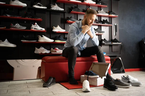 Cheerful man chooses shoes in a sports shop while sitting in fitting room of modern sports store