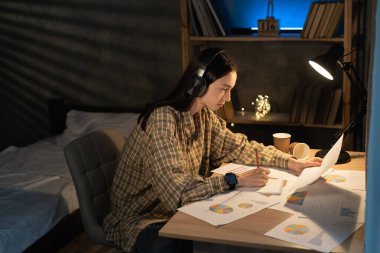 Proje üzerinde çalışan Asyalı bir kız öğrenci, finansal belgeleri kontrol ediyor, gece geç saatlerde masada oturuyor, diyagramlı evraklara bakıyor, faturaları hesaplıyor.