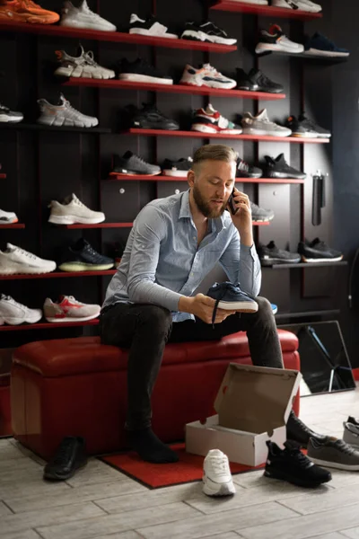 A man in a shoe store choosing sports sneakers, a buyer is talking on the phone trying on a pair of shoes, buying shoes in a shop, men\'s shopping concept