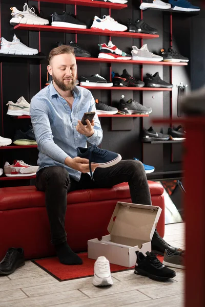 Young man choosing sneakers at sportswear shop, trying on shoes in the fitting room of a store, scanning a pair of sneakers with a smartphone