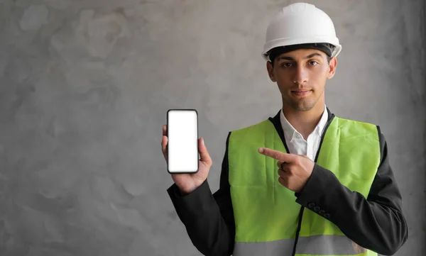 smiling Arabic male engineer or builder in white helmet and overall showing smartphone with empty screen over grey background. profession, construction and building concept