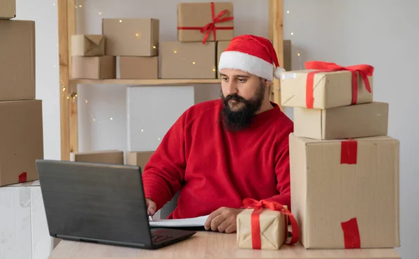 Indian small and medium business freelance Santa working in home office using computer, online marketing packaging box and gifts for Christmas delivery, SME e-commerce telemarketing concept.