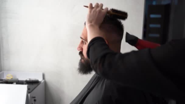 理容室でドライヤー付きの美容師さんに手入れをされている若い髭男の姿を間近で見ることができます 男性の髪をカットマスター 美容師は若い男のための髪型を作ります すでにいい — ストック動画